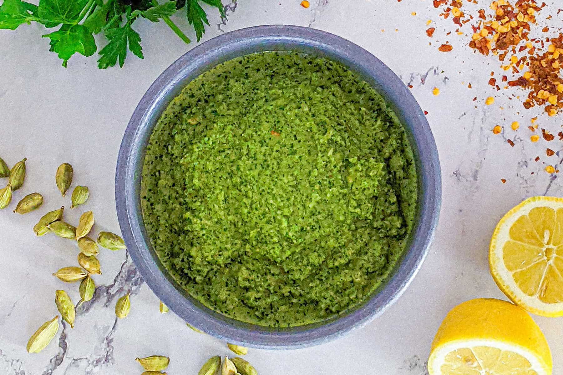 Zhug sauce in a bowl surrounded by lemons, chili pepper flakes, fresh parsley and cardamom pods.