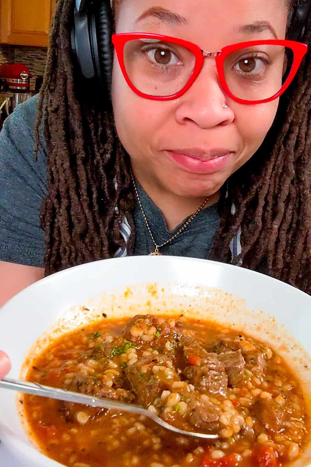 Chef Maika proudly holds a vibrant, hearty spicy beef barley stew in a wide bowl in a cozy kitchen.