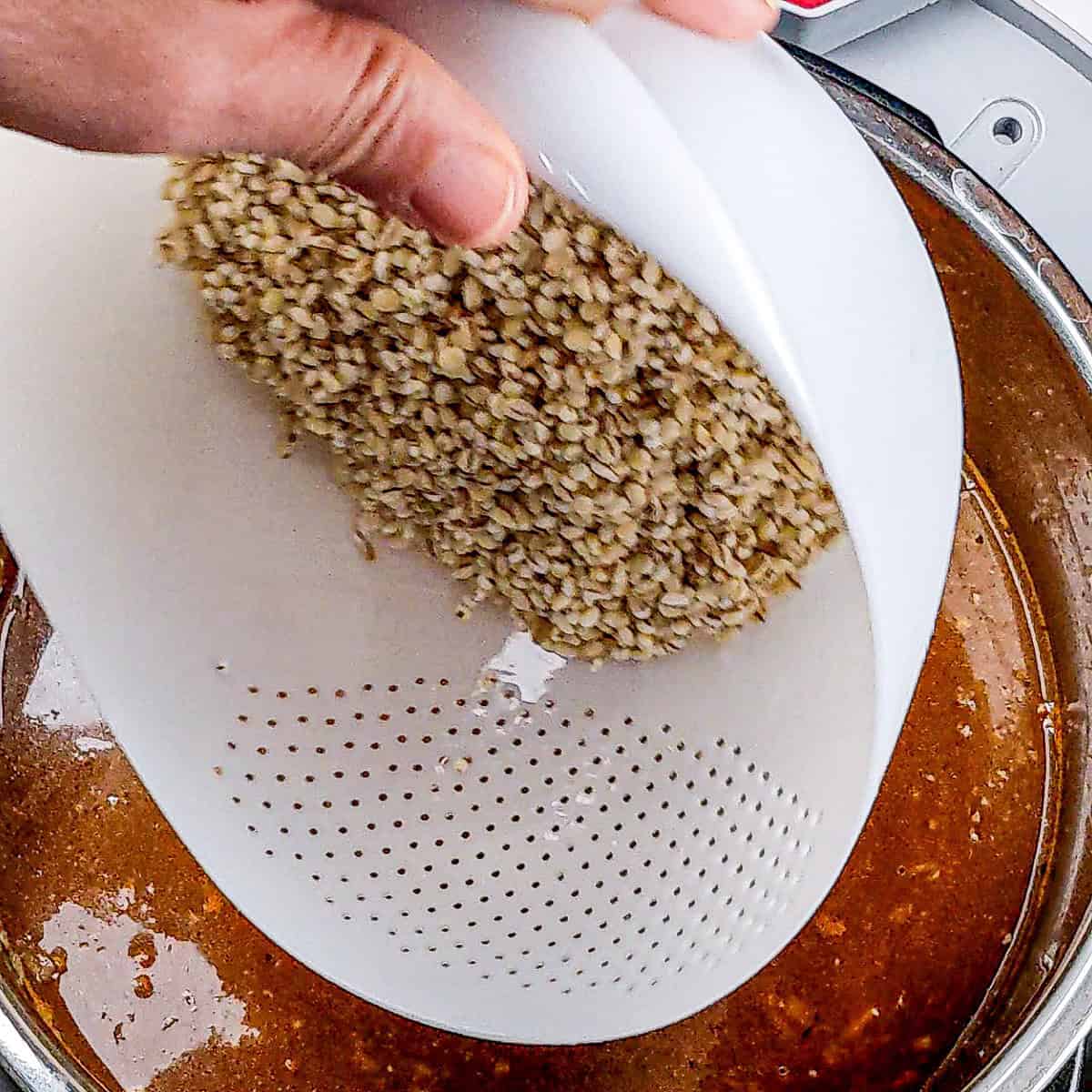 Hand pouring grains into simmering spicy beef barley stew for a flavorful meal.