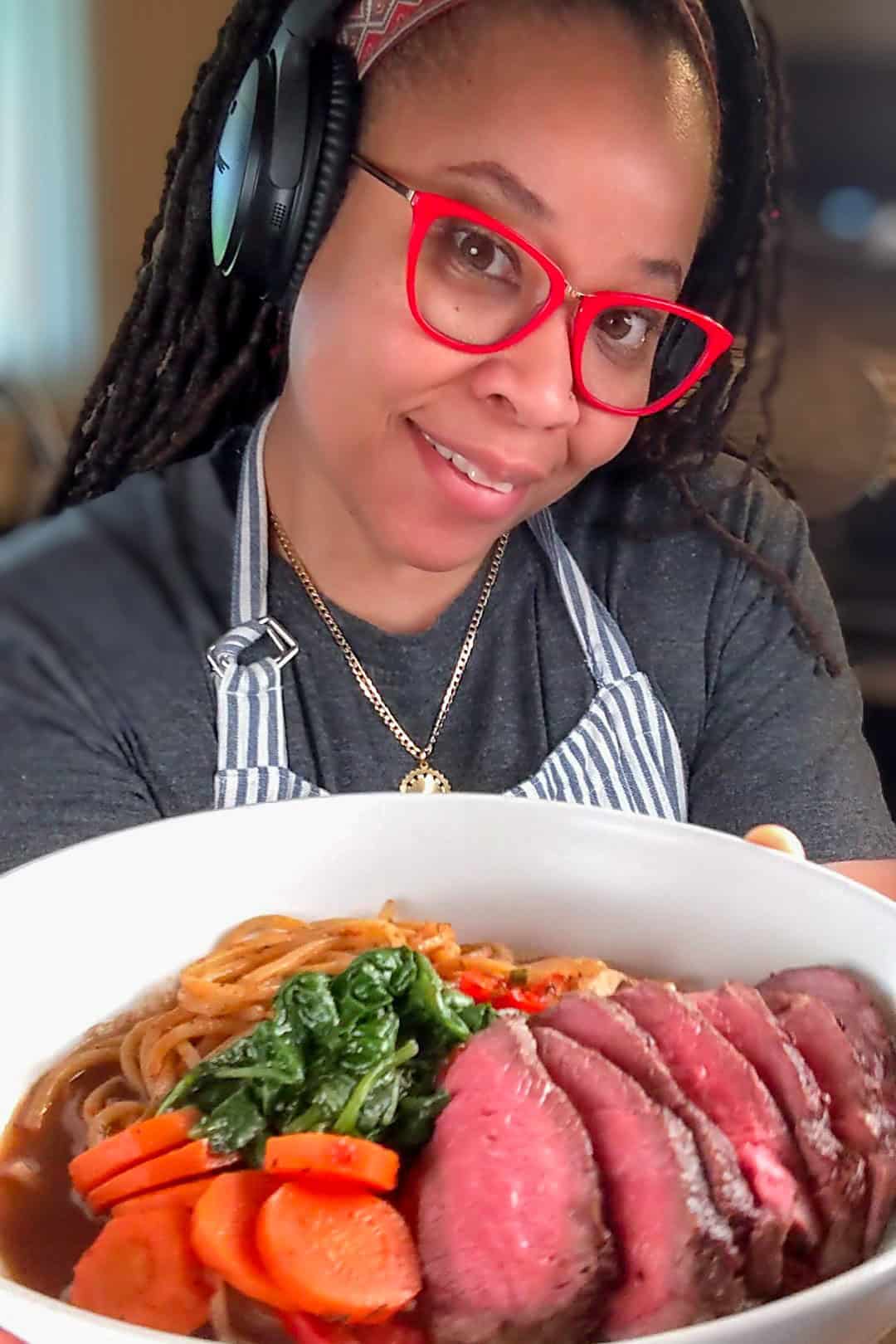 Smiling Chef Maika presents vibrant Caribbean beef noodle soup in a cozy kitchen setting.