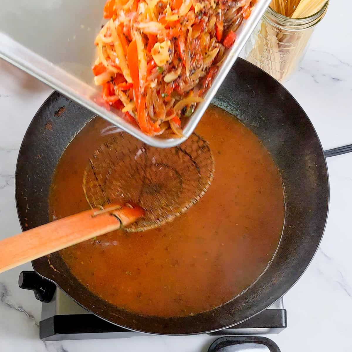 Sautéed vegetables added to hearty Caribbean beef noodle soup in a cast iron skillet.