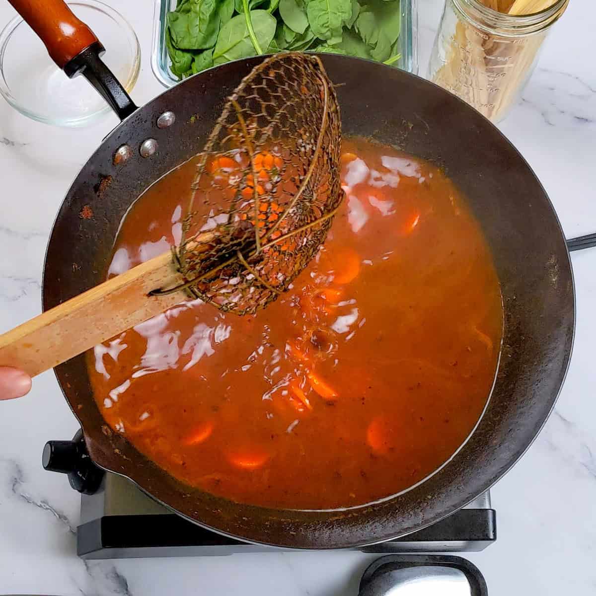 Wooden spoon stirring Caribbean beef noodle soup in a dark pot on a marble countertop.