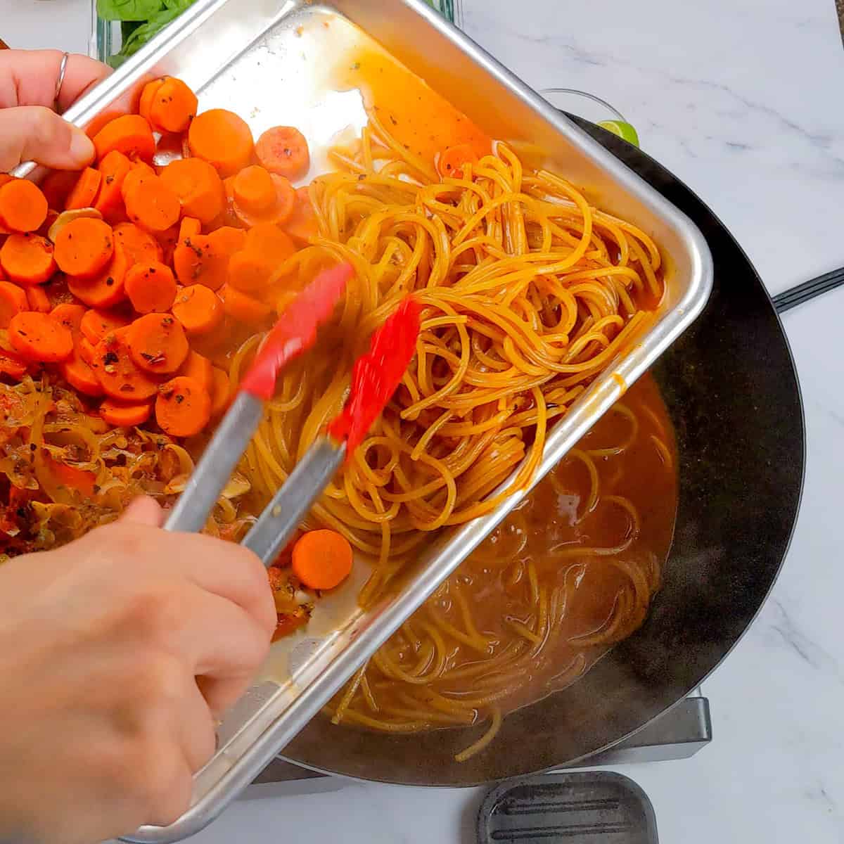 Hands using tongs to add vibrant ingredients to Caribbean beef noodle soup in skillet.