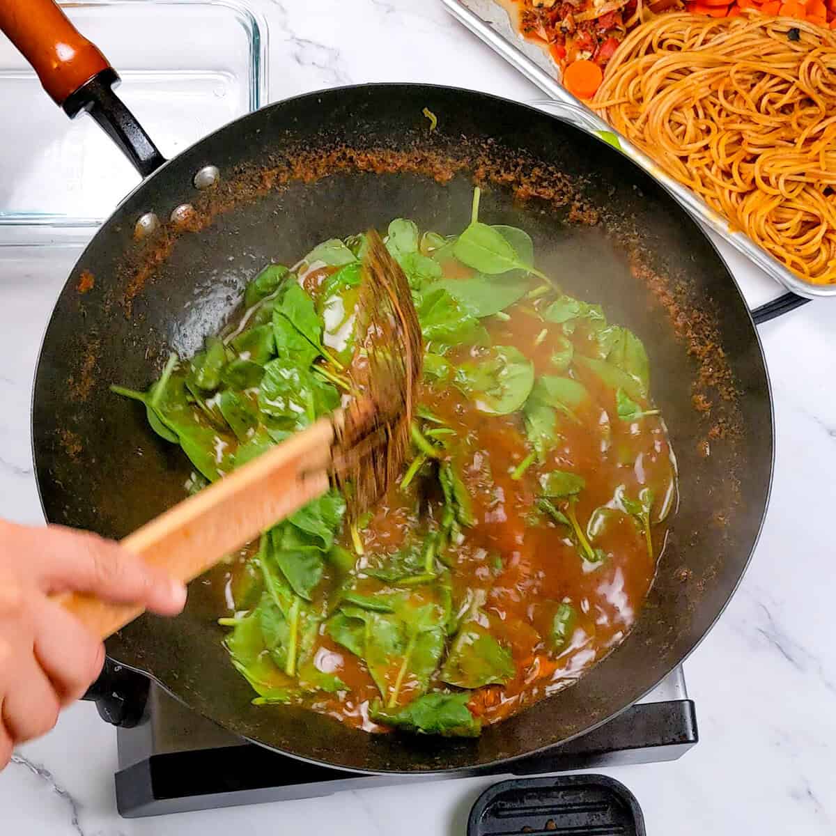 Sautéed spinach in wok complements Caribbean beef noodle soup preparation for a flavorful meal.