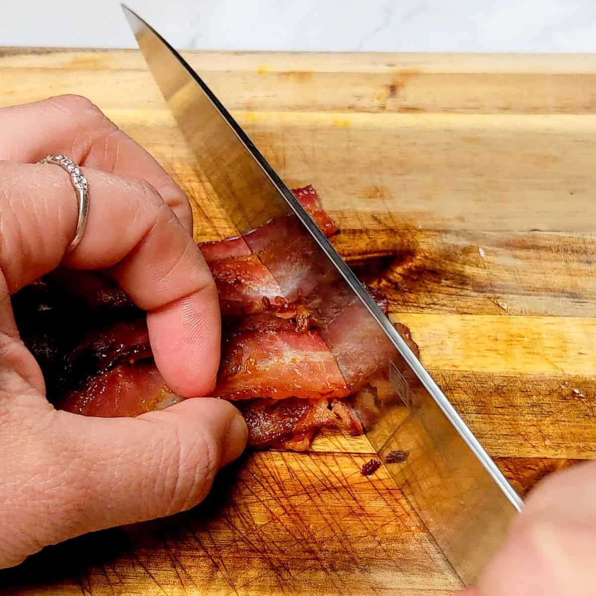 Close-up of a hand slicing cooked bacon on a wooden cutting board with a sharp knife.