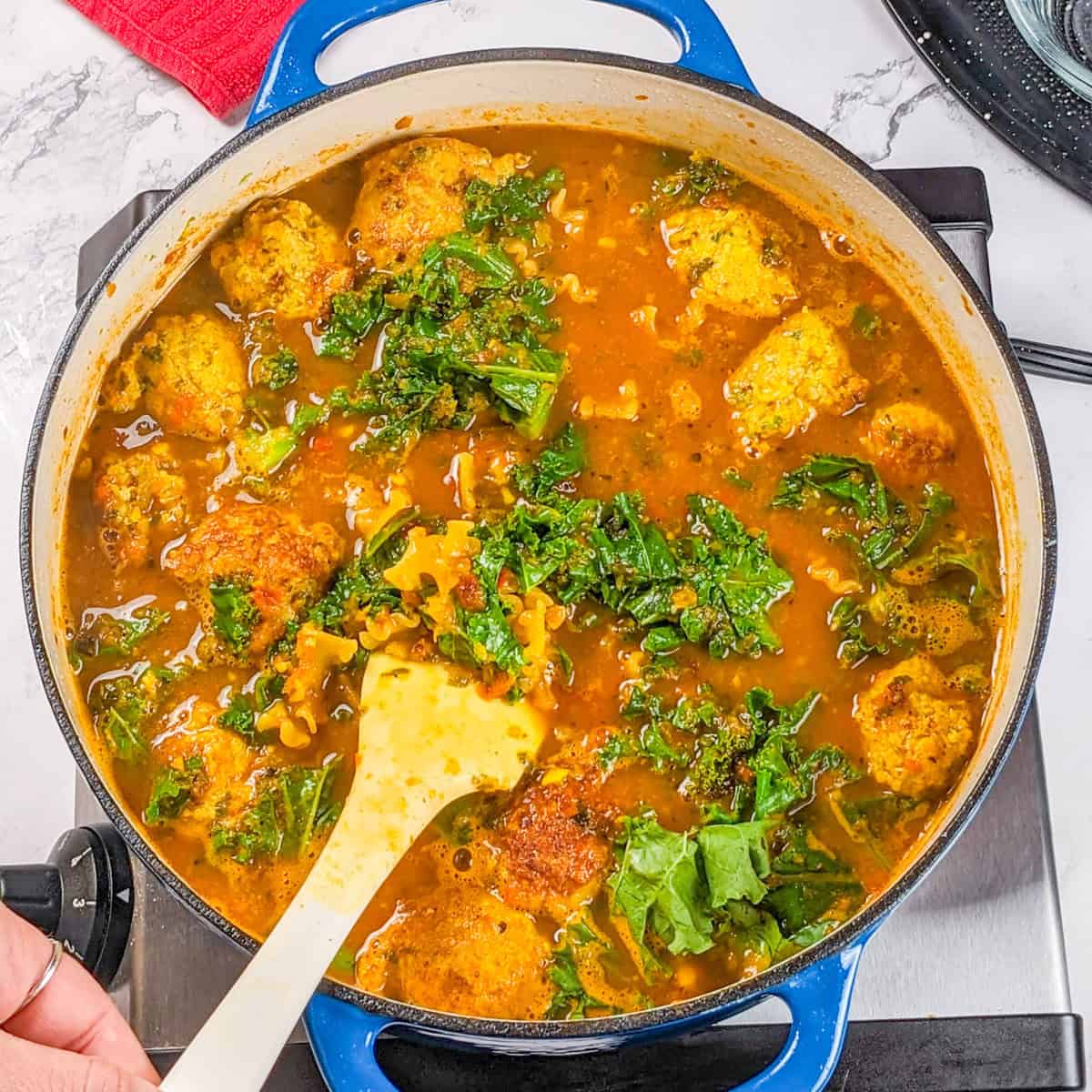 Hearty curry turkey meatball soup with golden noodle and vibrant kale in a blue pot.