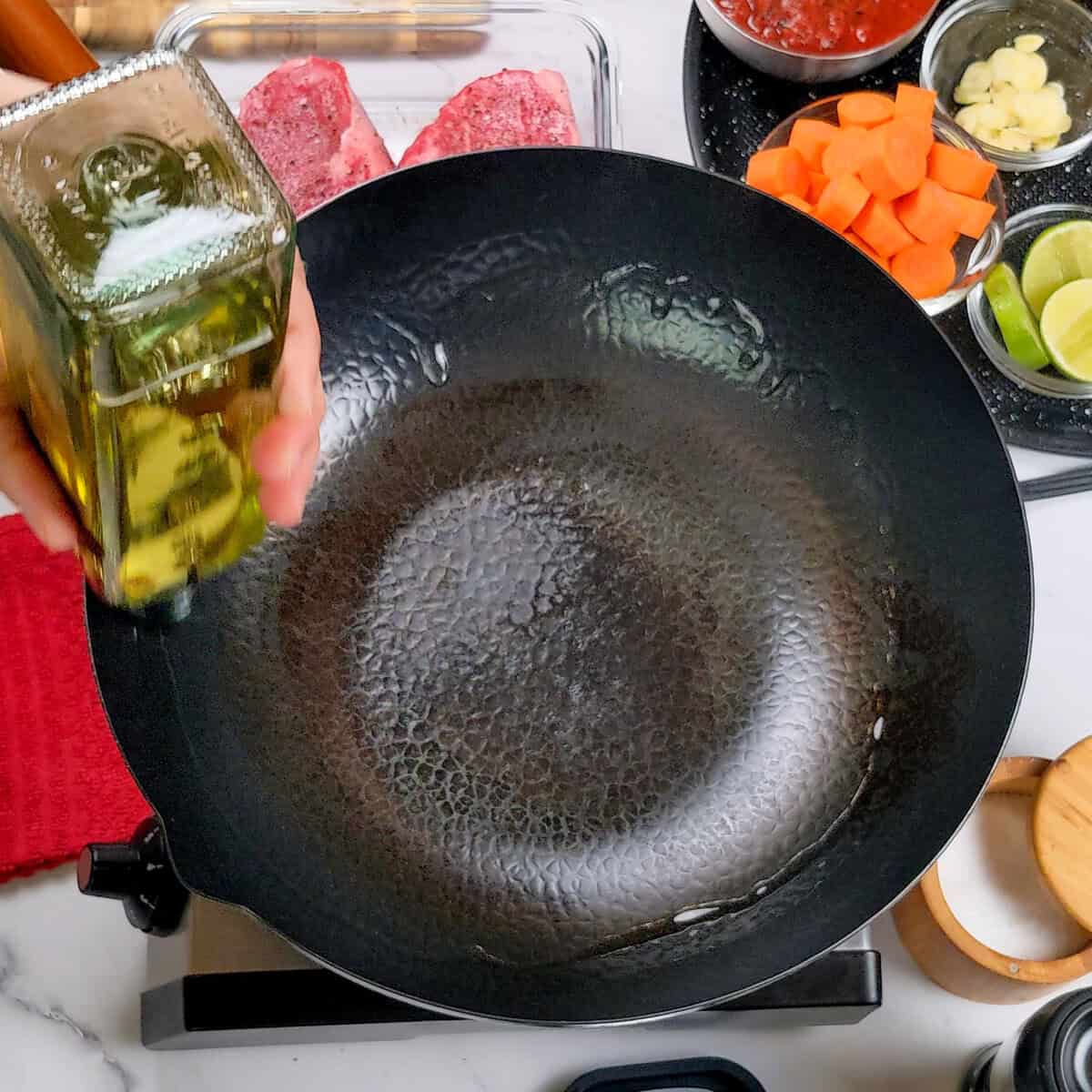 Chef Maika pouring oil into a glossy wok around the rim surrounded by fresh ingredients for a stir-fry.