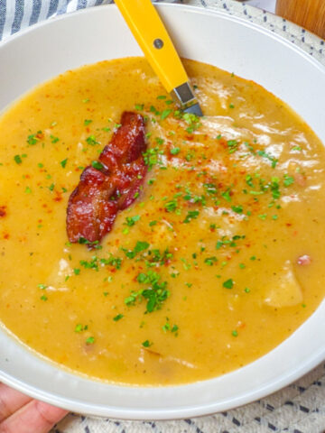 wide rim bowl filled with Creamy spicy bacon potato leek chowder that's garnished with a half of slice of crispy bacon and chopped parsley then dusted with smoked paprika held in hand.