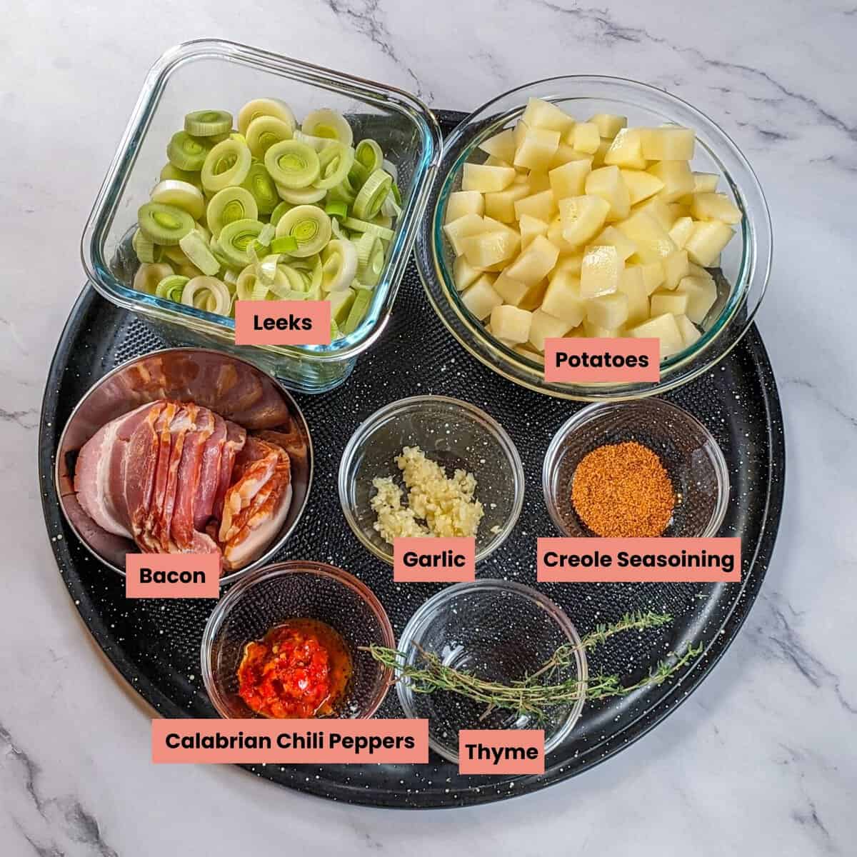 Ingredients for spicy bacon potato leek chowder arranged on a dark platter against a marble countertop.