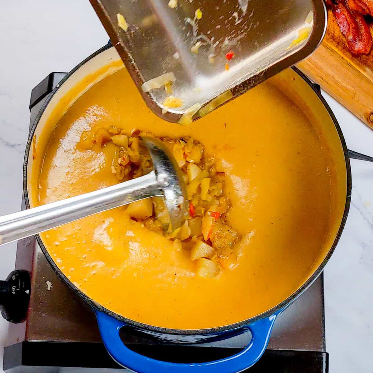 Blending creamy bacon potato leek chowder in a vibrant enameled cast-iron dutch oven on the stovetop with a ladle adding back the potato mixture.