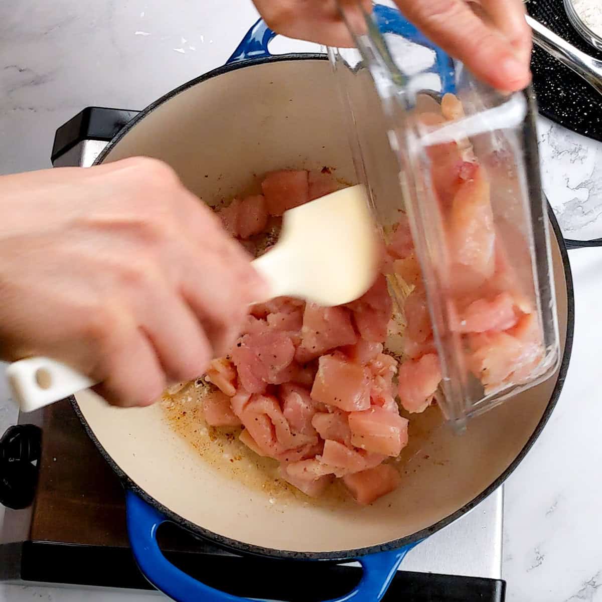 raw diced chicken being poured into a lodge cast iron enameled dutch oven.