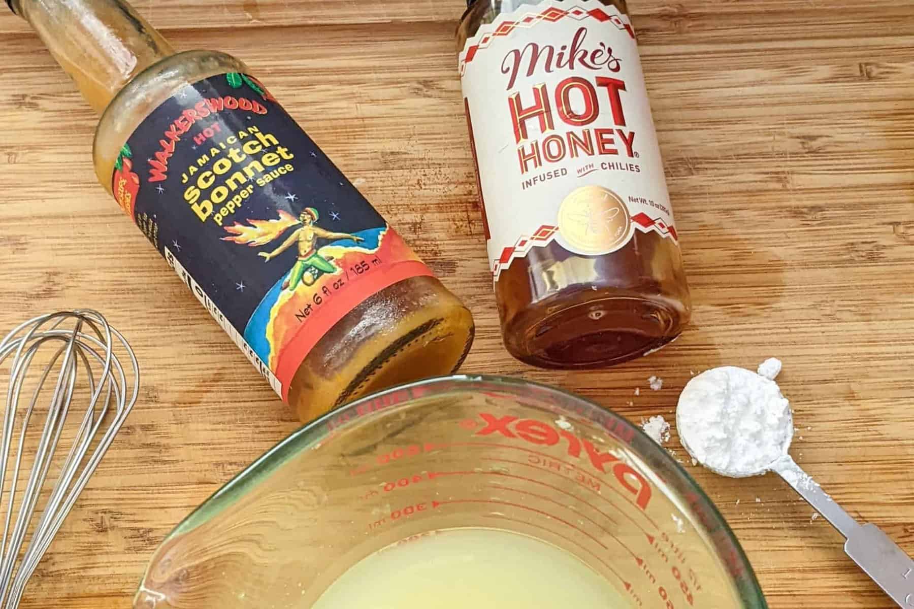 chicken broth in a glass measuring cup, next to mike's hot honey and walkerwood's scotch bonnet pepper sauce.