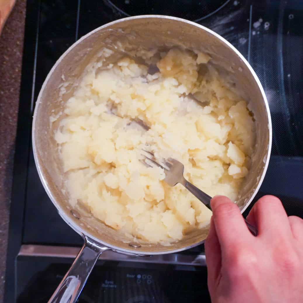 Mashed potatoes in a saucepan being mixed with a fork on a modern stovetop.