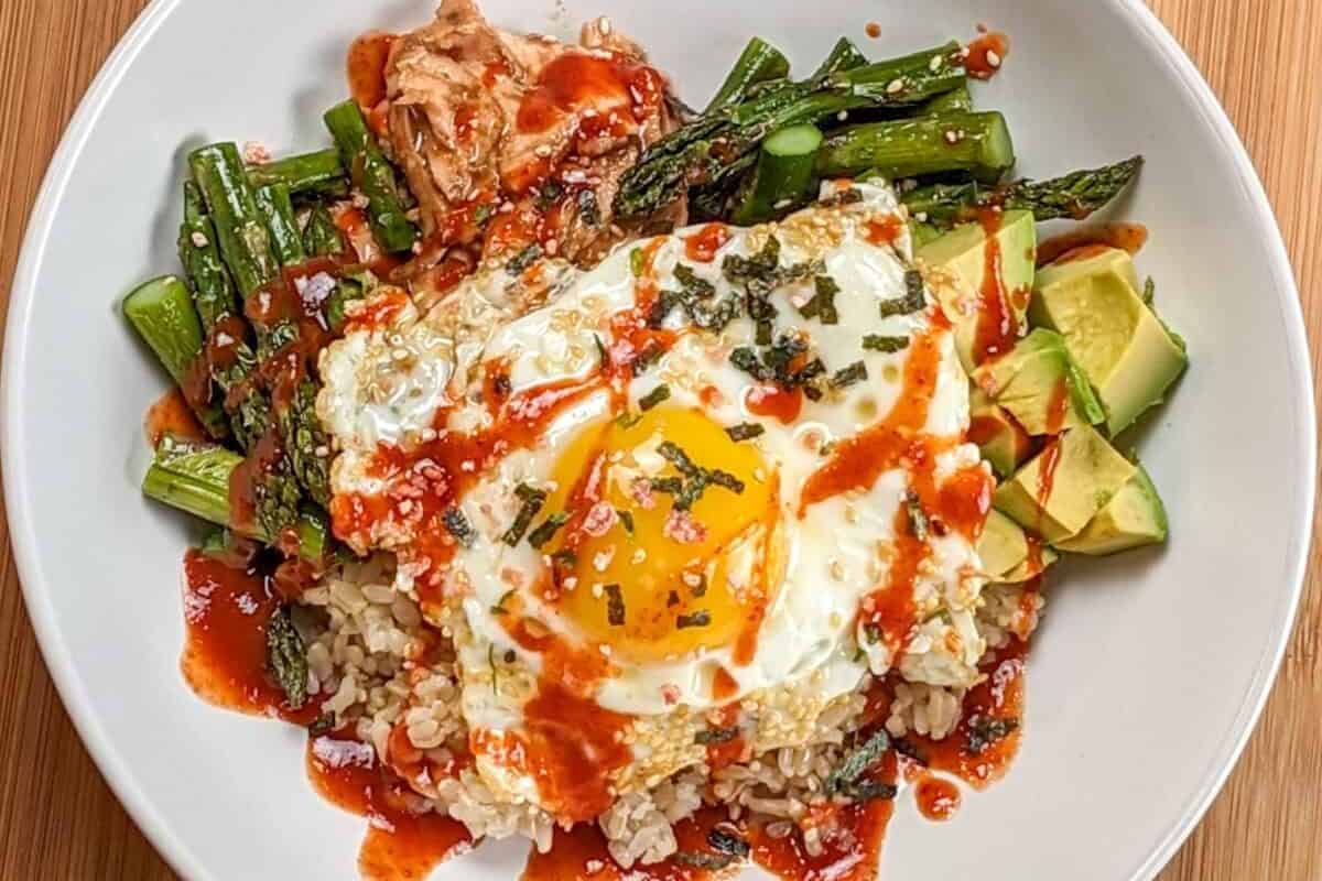 the Easy Sheet Pan Spicy Miso Salmon Rice Bowl in a wide rim bowl drizzled with siracha, sesame oil and salmon furikake seasoning.