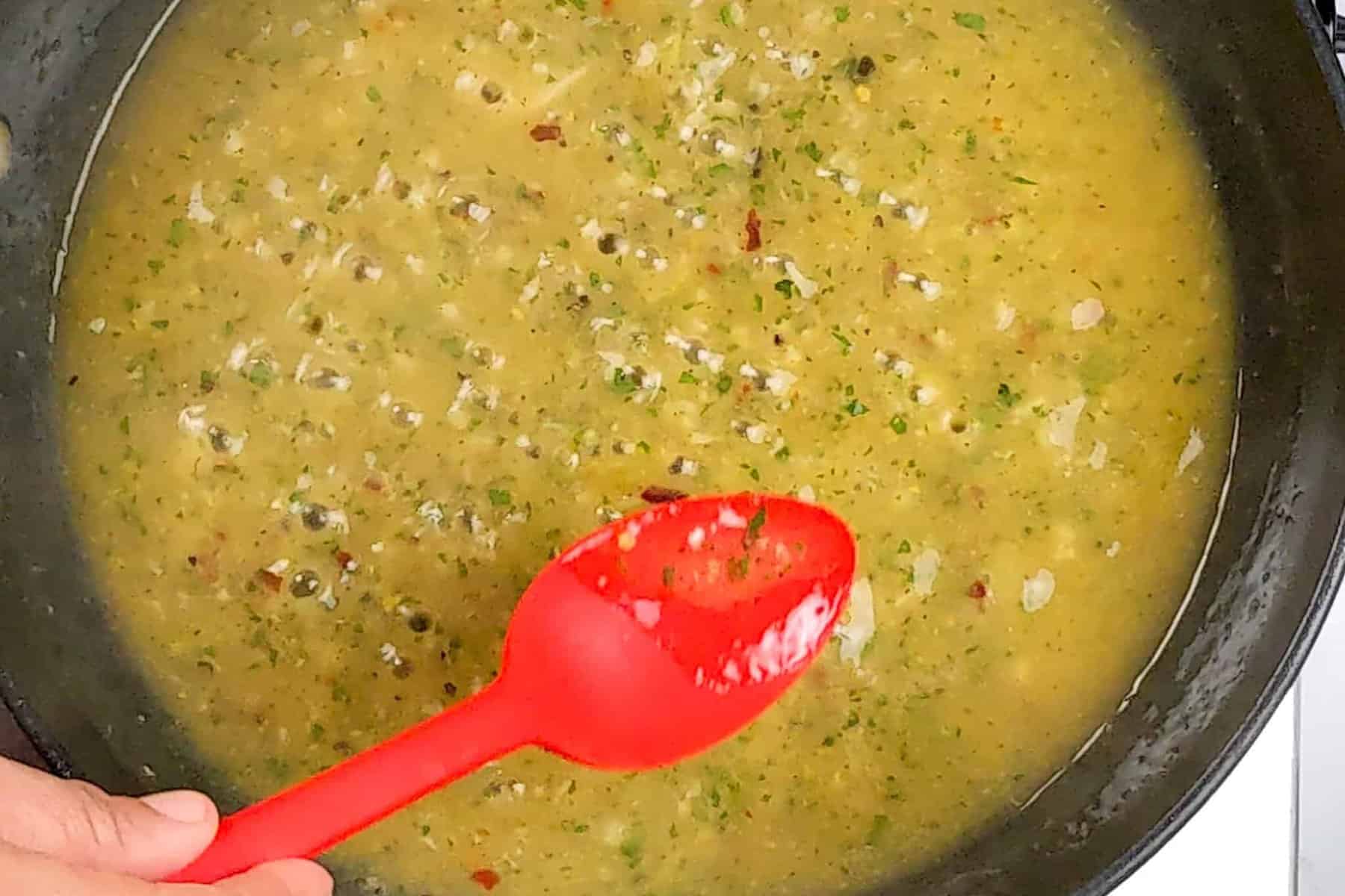 the wet lemon pepper sauce reducing with small bubbles in a non-stick frying pan being tasted with a silicone red spoon