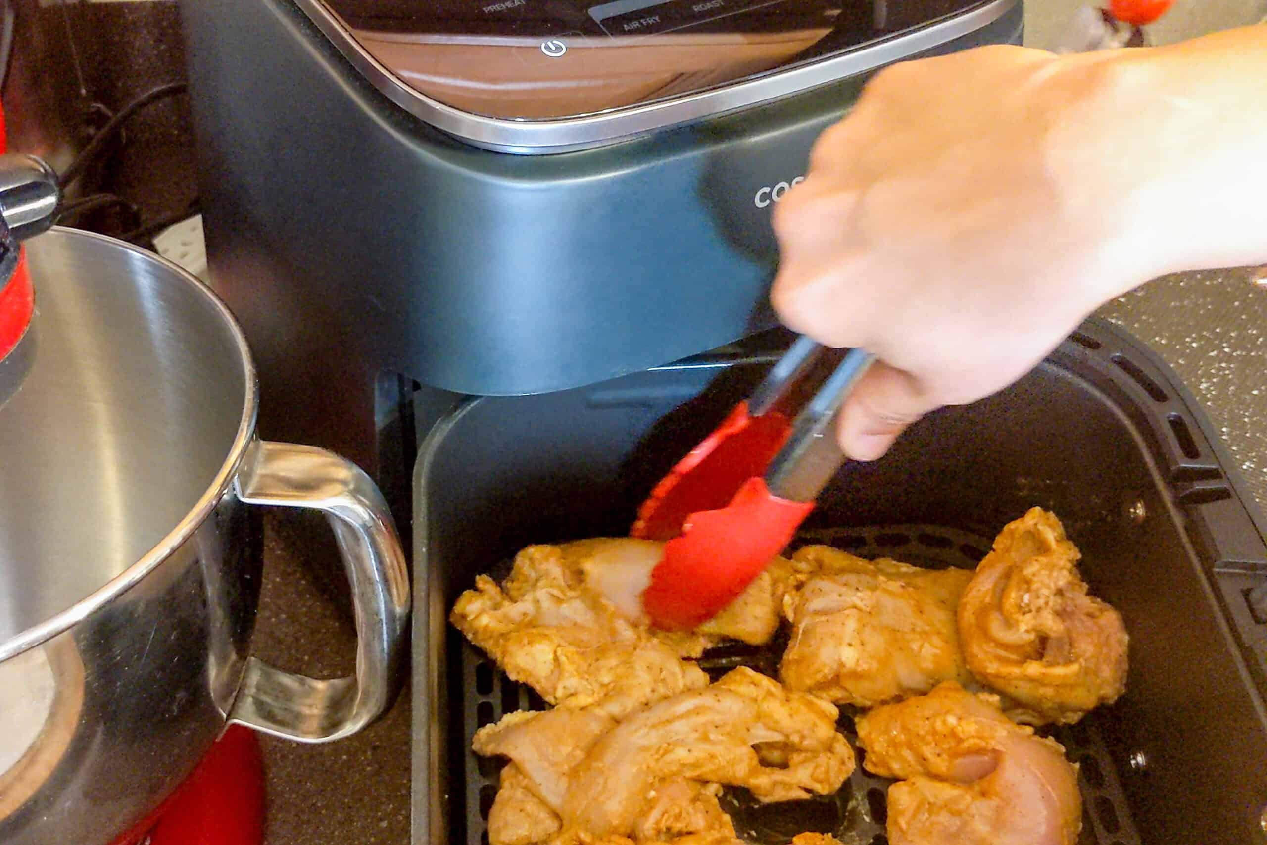 marinated chicken thighs being placed in the air fryer basket with silicone tipped tongs.