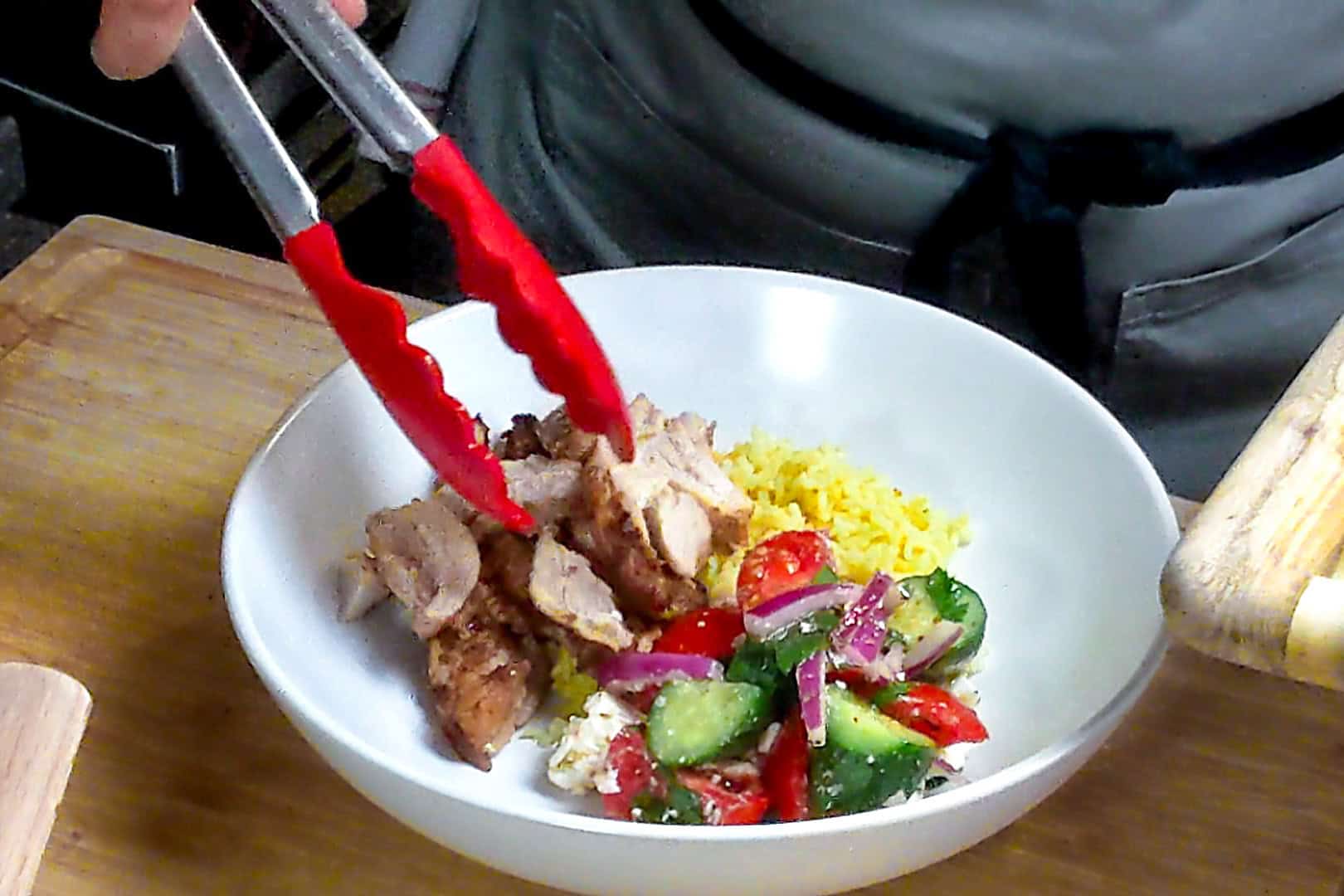 the sliced beituti chicken shawarma being plated in a rice bowl with tongs.