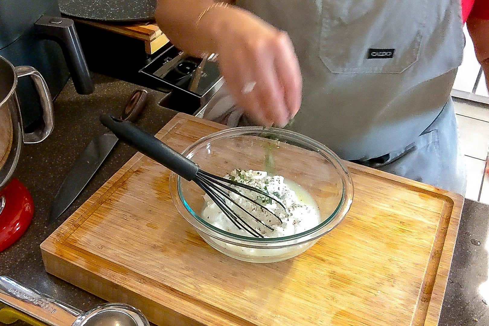 herbs being sprinkled into the greek yogurt mixture in a glass mixing bowl with a wihisk.