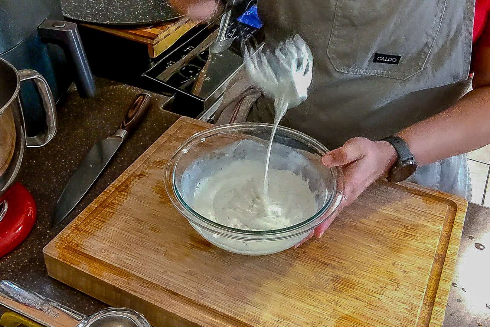 chef maika testing the consistency of the lemon greek yogurt dressing with a silicone whish in a glass mixing bowl.