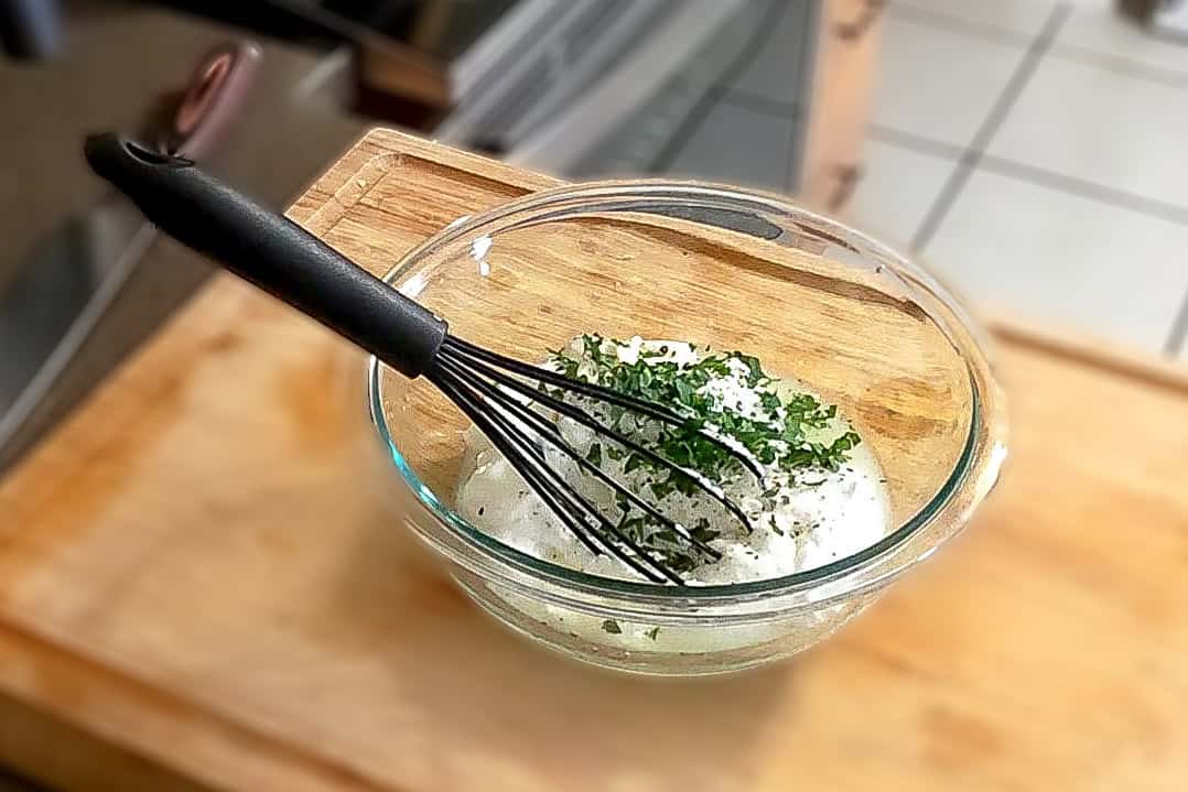 lemon greek yogurt mixture sprinkled with fresh chopped parsley sitting in a glass mixing bowl with a silicone whisk.