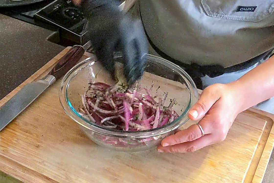 ingredients for the sumac onions in a glass mixing bowl being mixed with a gloved hand.