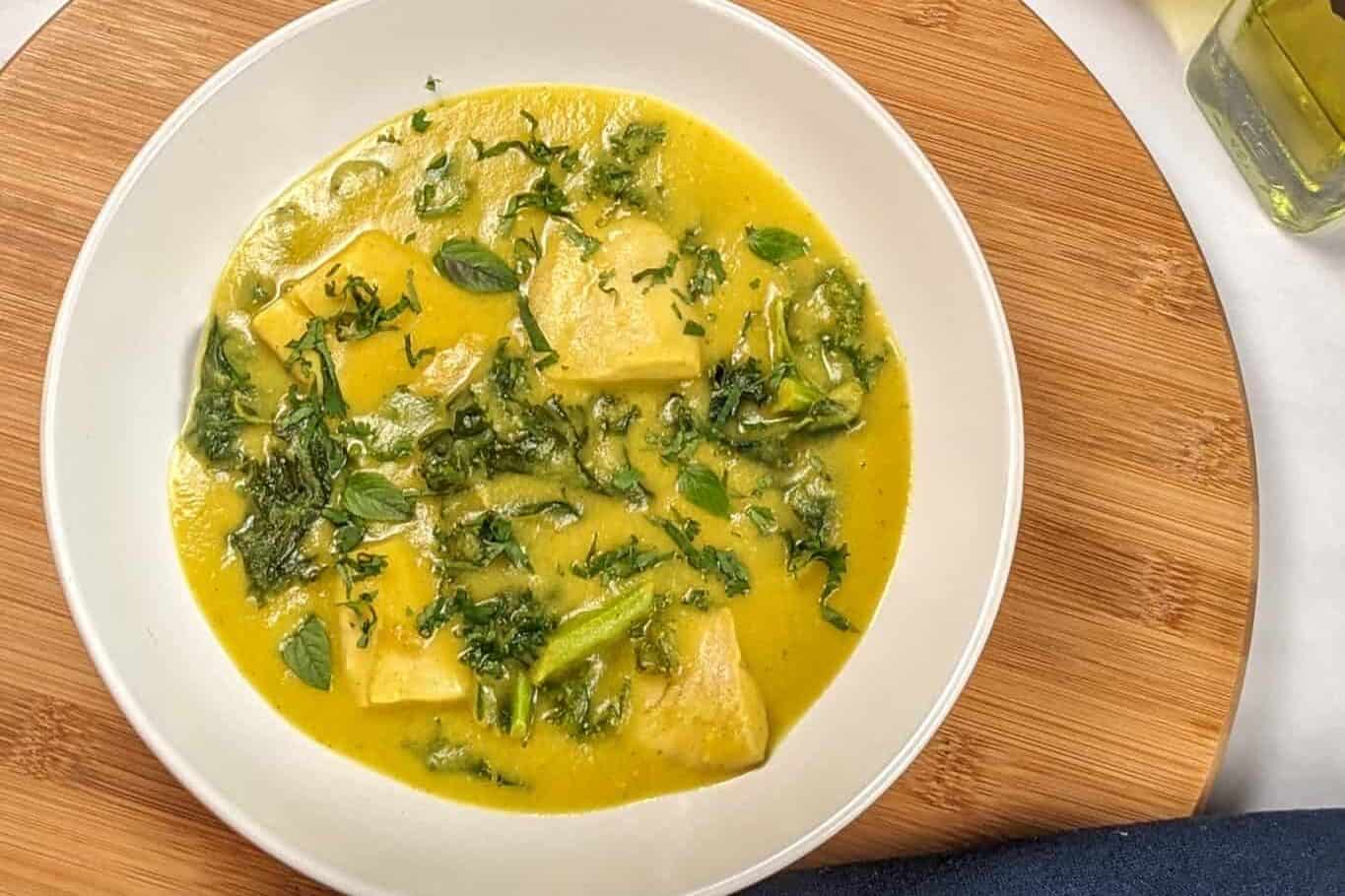 top view of the Best Fragrant Creamy Potato Coconut Curry Fish Soup in a wide rim soup bowl on a wooden lazy susan next to a kitchen towel with the dutch oven with the rest of the soup on the counter next to a salt cellar